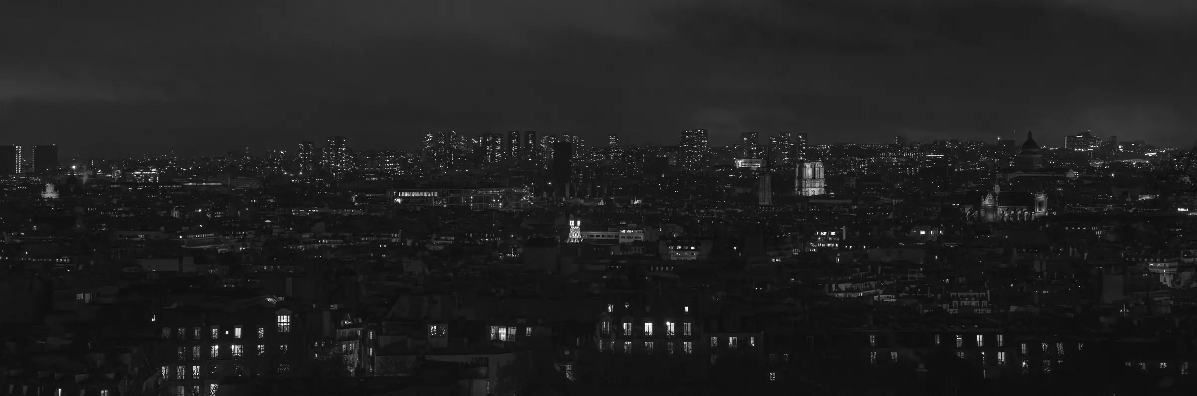 Paris la nuit, vu de haut : une longue bande de lumières de la ville sous un ciel bas et sombre. Noir et blanc.
